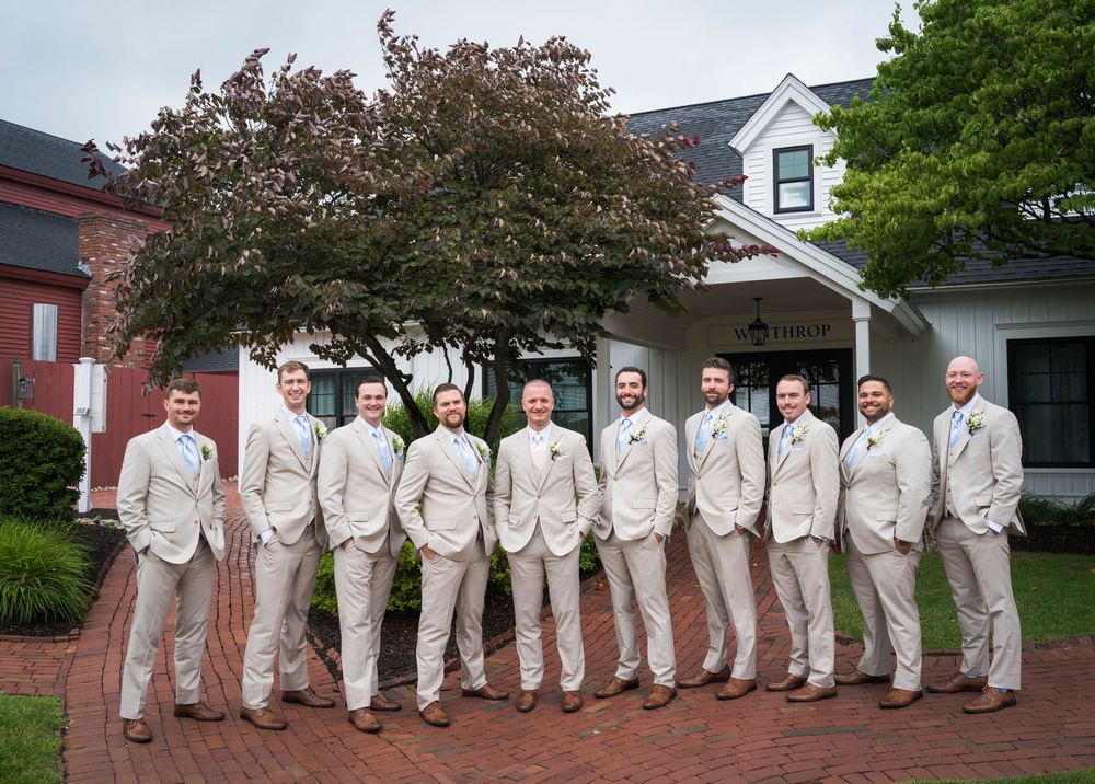 Groom and groomsmen pose in matching tan suits outside elegant white building on wedding day.