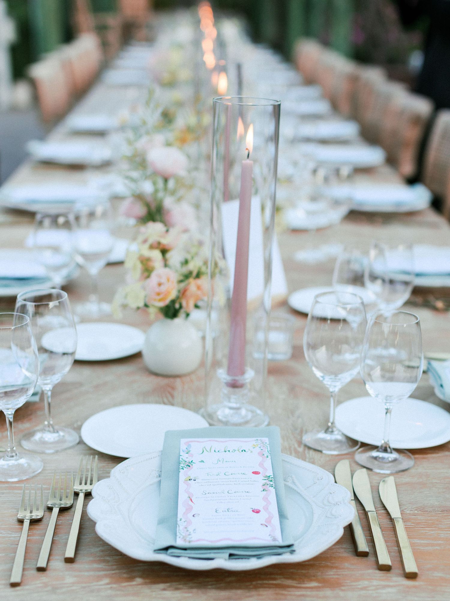Elegant wedding table setting with white plates, pink candles, and delicate floral arrangements on a rustic wooden surface.