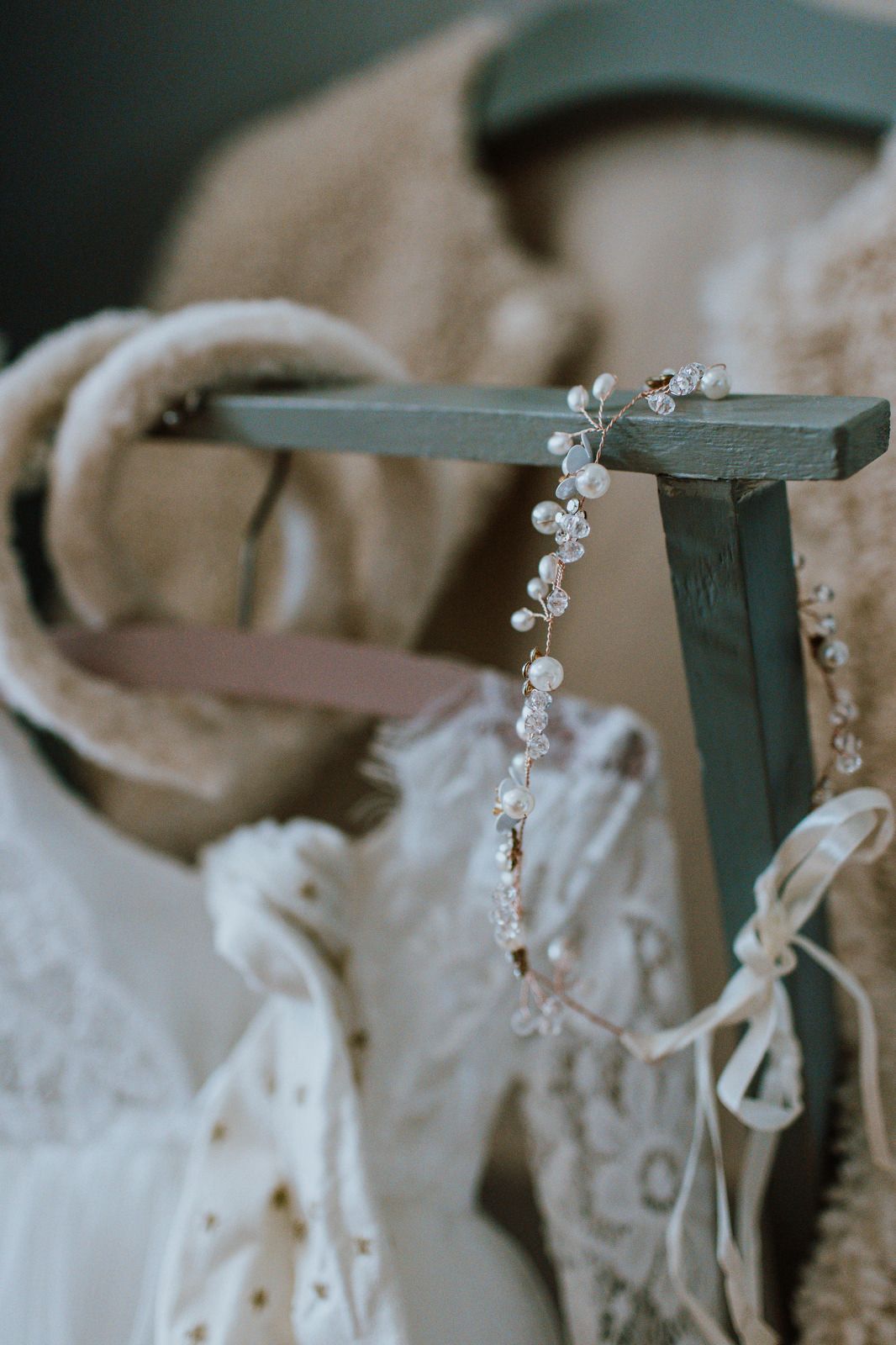 A delicate pearl and crystal bridal headpiece hangs on a green stand with white lace fabric in the background.