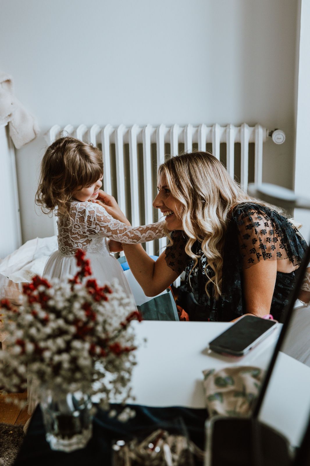 Wedding party preparations show women getting ready together in a bright room with white radiators and floral arrangements.