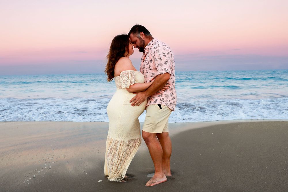 Expecting couple in a romantic embrace on a sandy beach at sunset, with gentle waves and a pink and purple sky behind them.