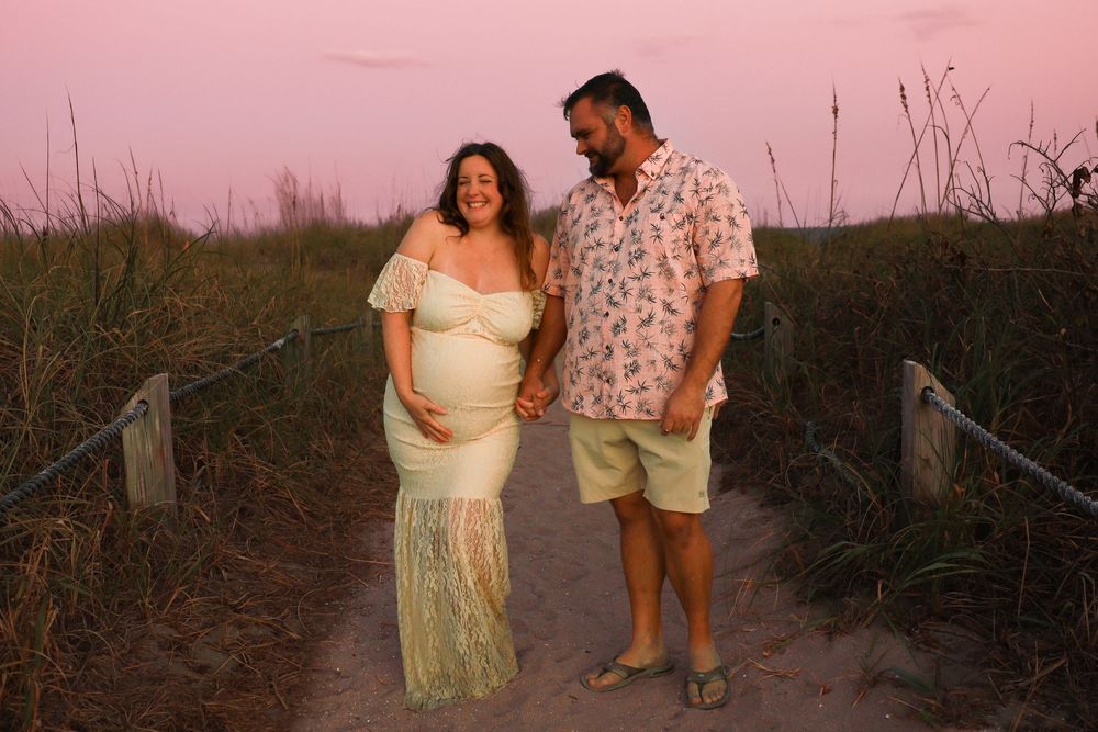 Expecting couple holds hands on a sandy beach path lined with dune grass during a pink sunset maternity photoshoot.