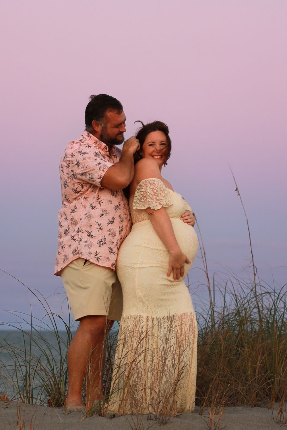 Smiling pregnant woman in a cream lace dress embraced by a bearded man in a floral shirt on a beach with sea oats at sunset.