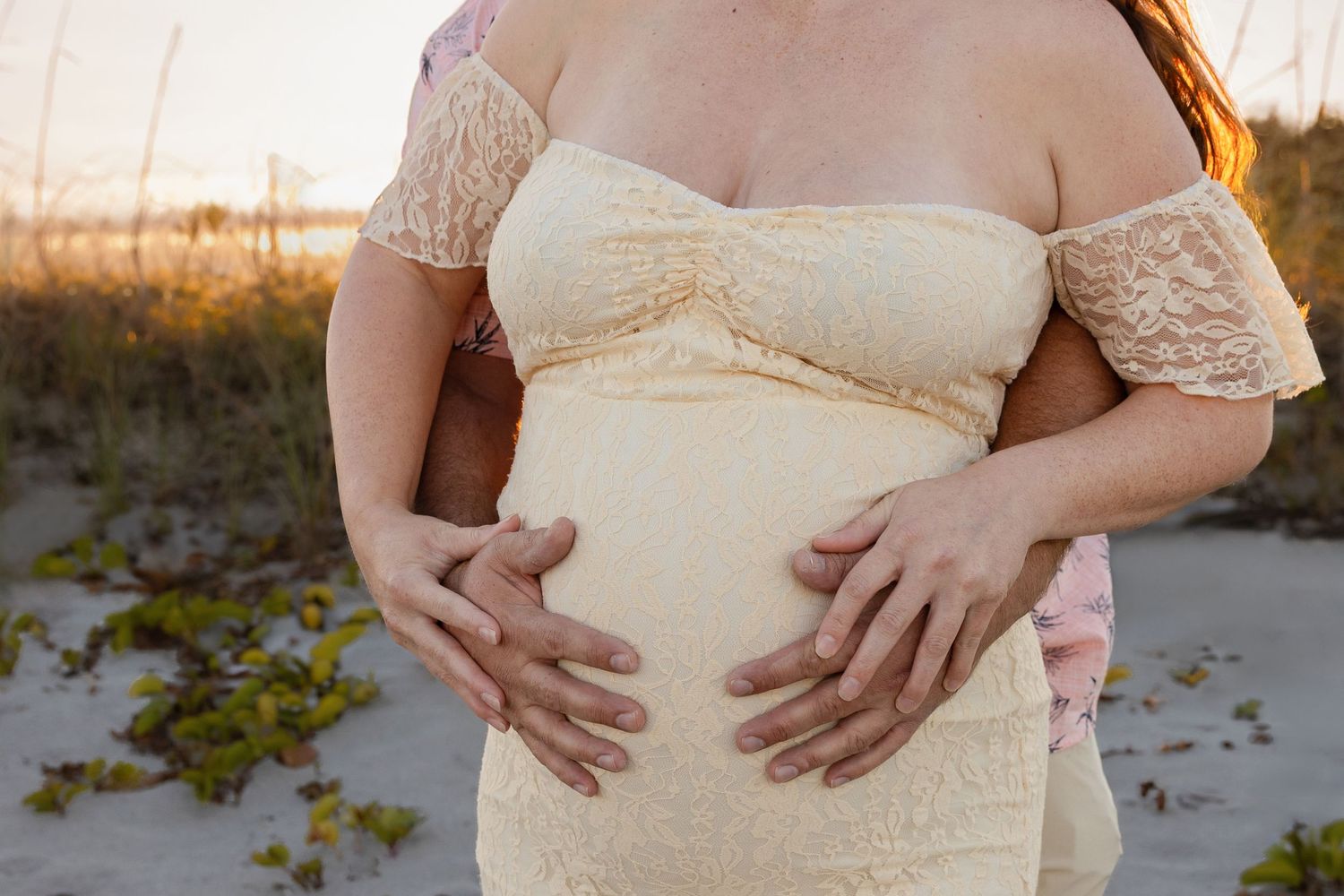 Couple cradles pregnant belly together in cream lace maternity dress outdoors.