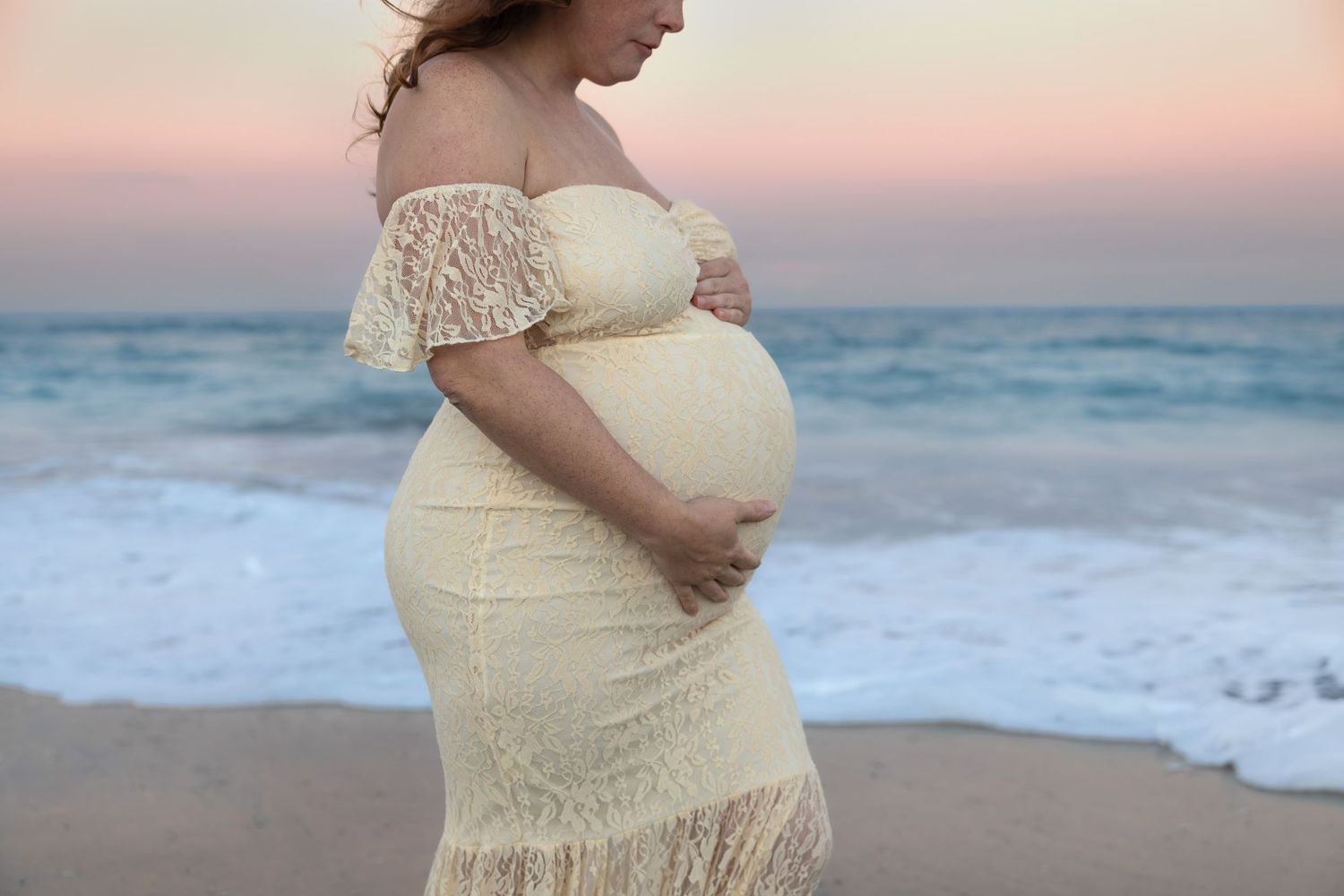 Close-up of pregnant woman in cream lace dress cradling belly on beach.