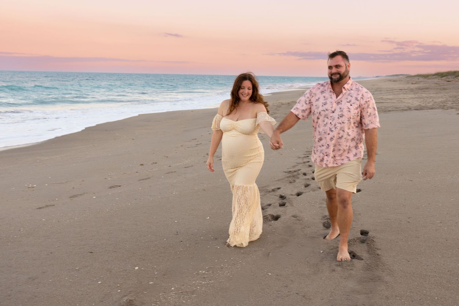 Couple holding hands walking on beach at sunset, woman in white maternity gown.