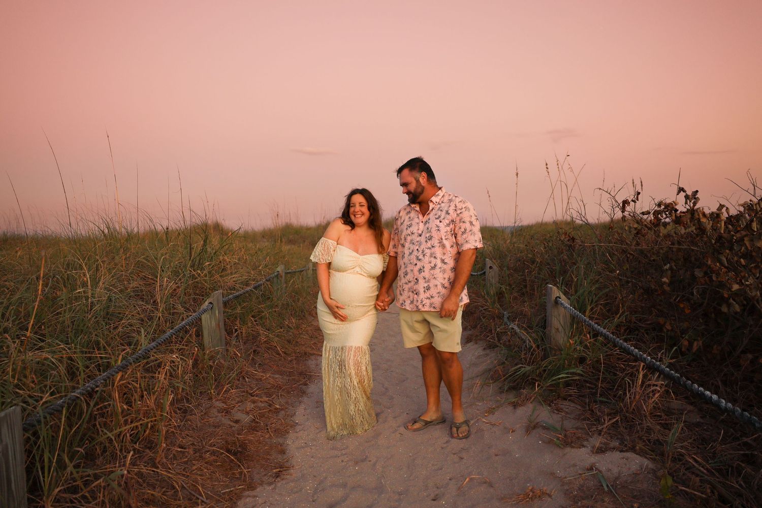 Expecting couple on sandy beach path at dusk, woman in white maternity dress.