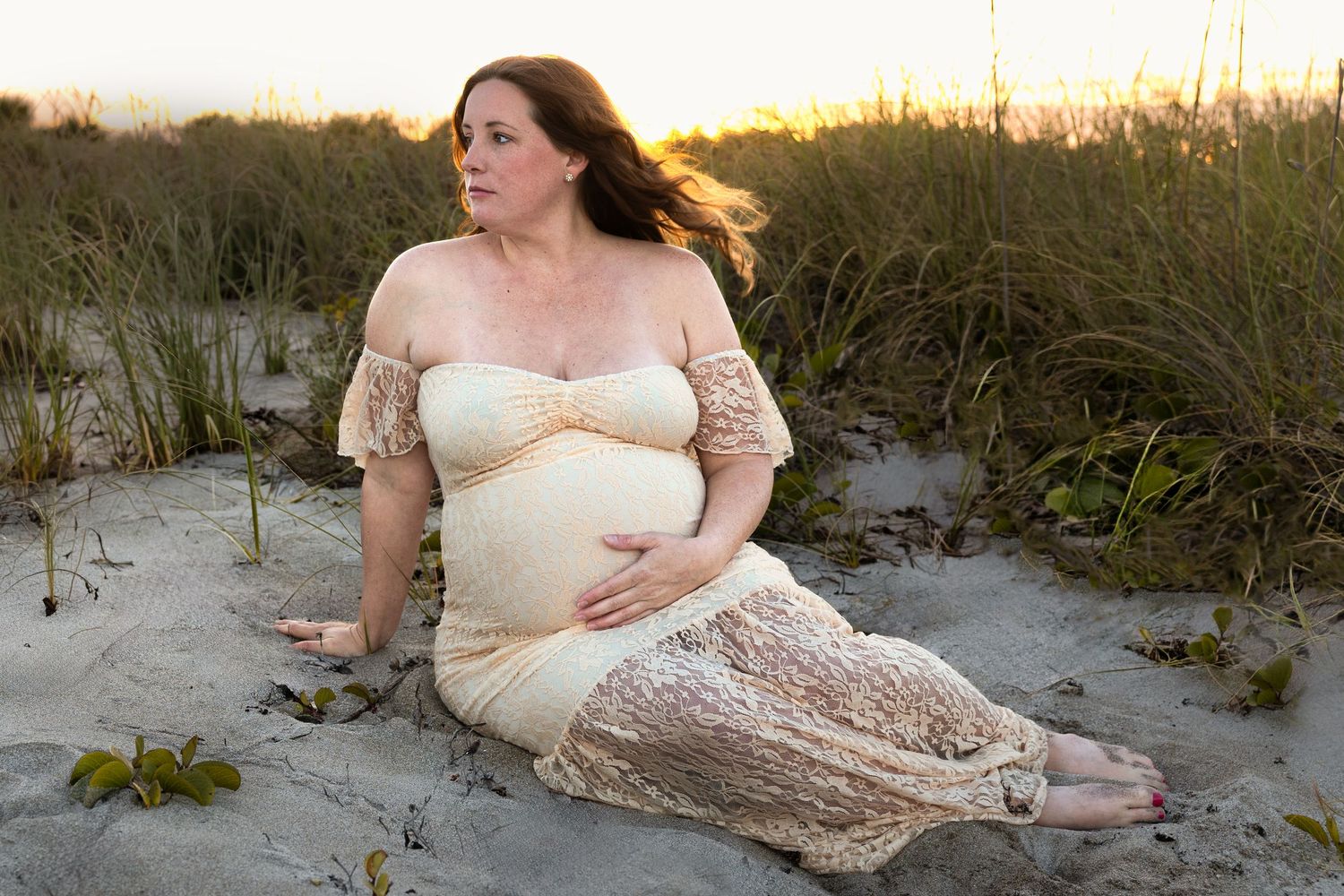 Pregnant woman in lace dress sitting on beach sand dunes at golden hour.