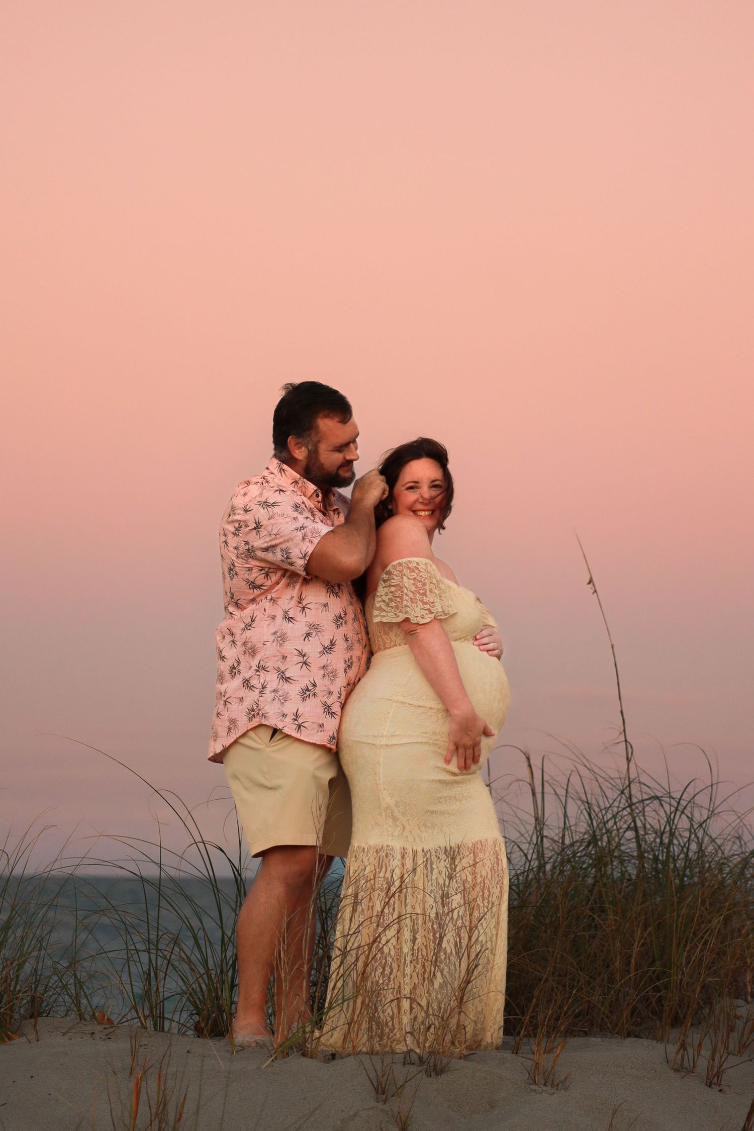 Couple embraces on sand dunes at pink sunset, woman in lace maternity gown.