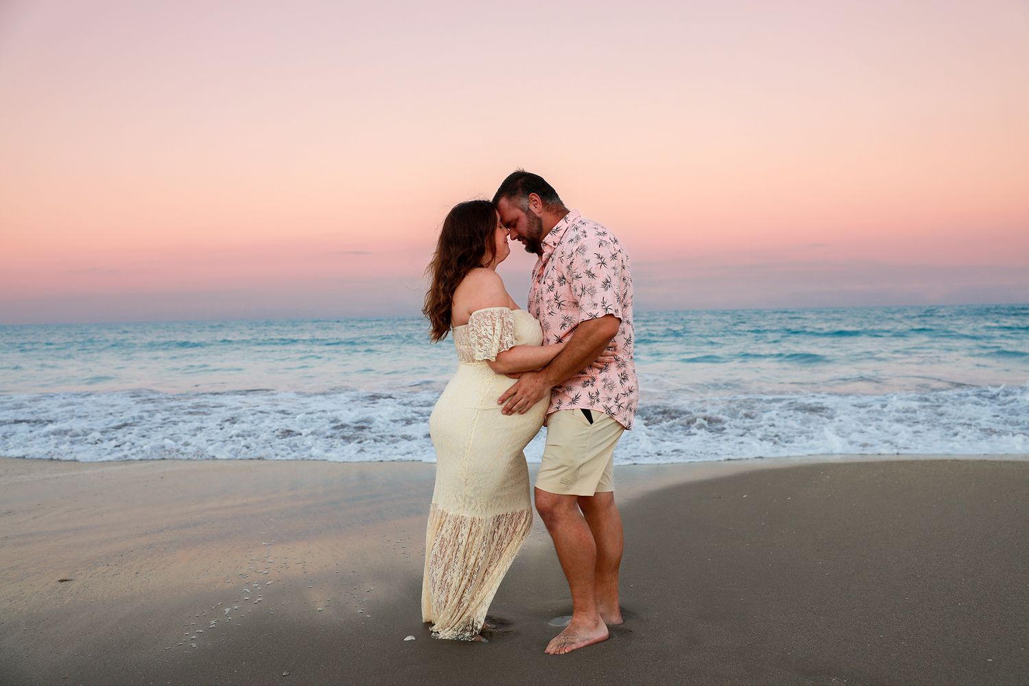 Pregnant couple embracing on beach at pink sunset, woman in white lace maternity gown.