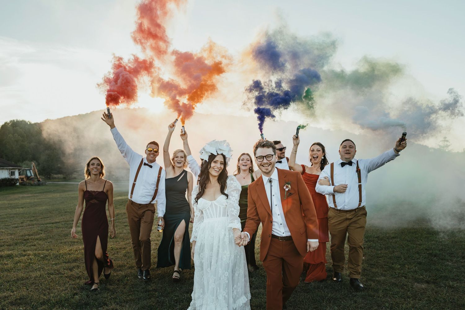 Wedding party celebrates outdoors with colorful smoke bombs in a grassy field at sunset.