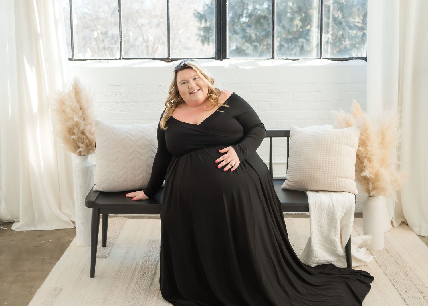 A person in a flowing black dress poses elegantly in a bright, sunlit room with white curtains and decor.
