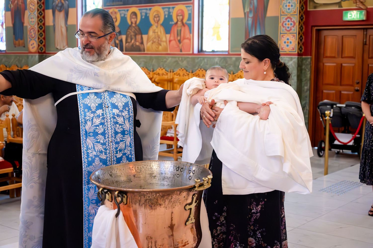 Orthodox priest performing a baby baptism ceremony at a church font, with a woman holding the infant in white christening attire.