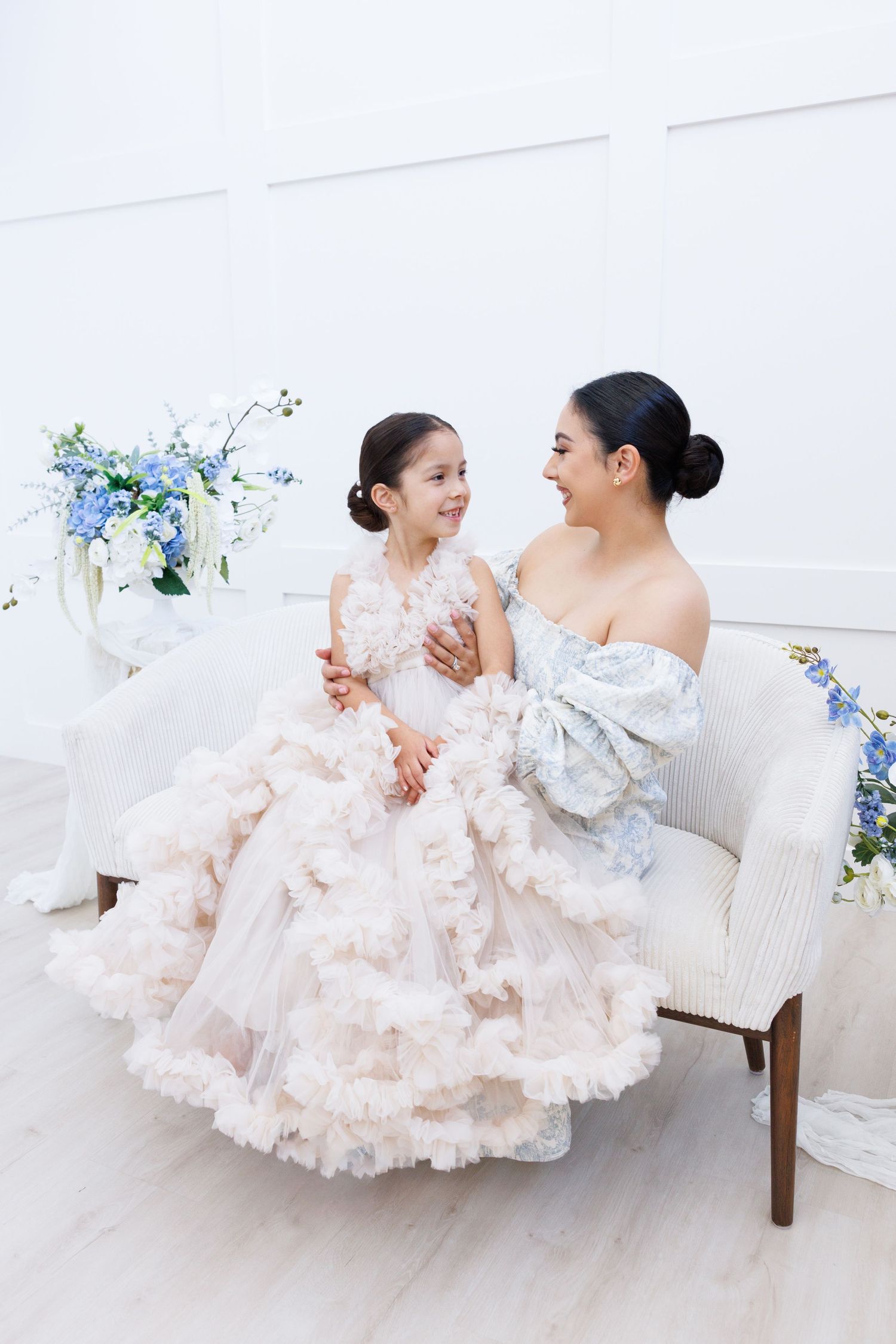 Mother and daughter in matching blue floral dresses sit together on a white couch in a bright room.