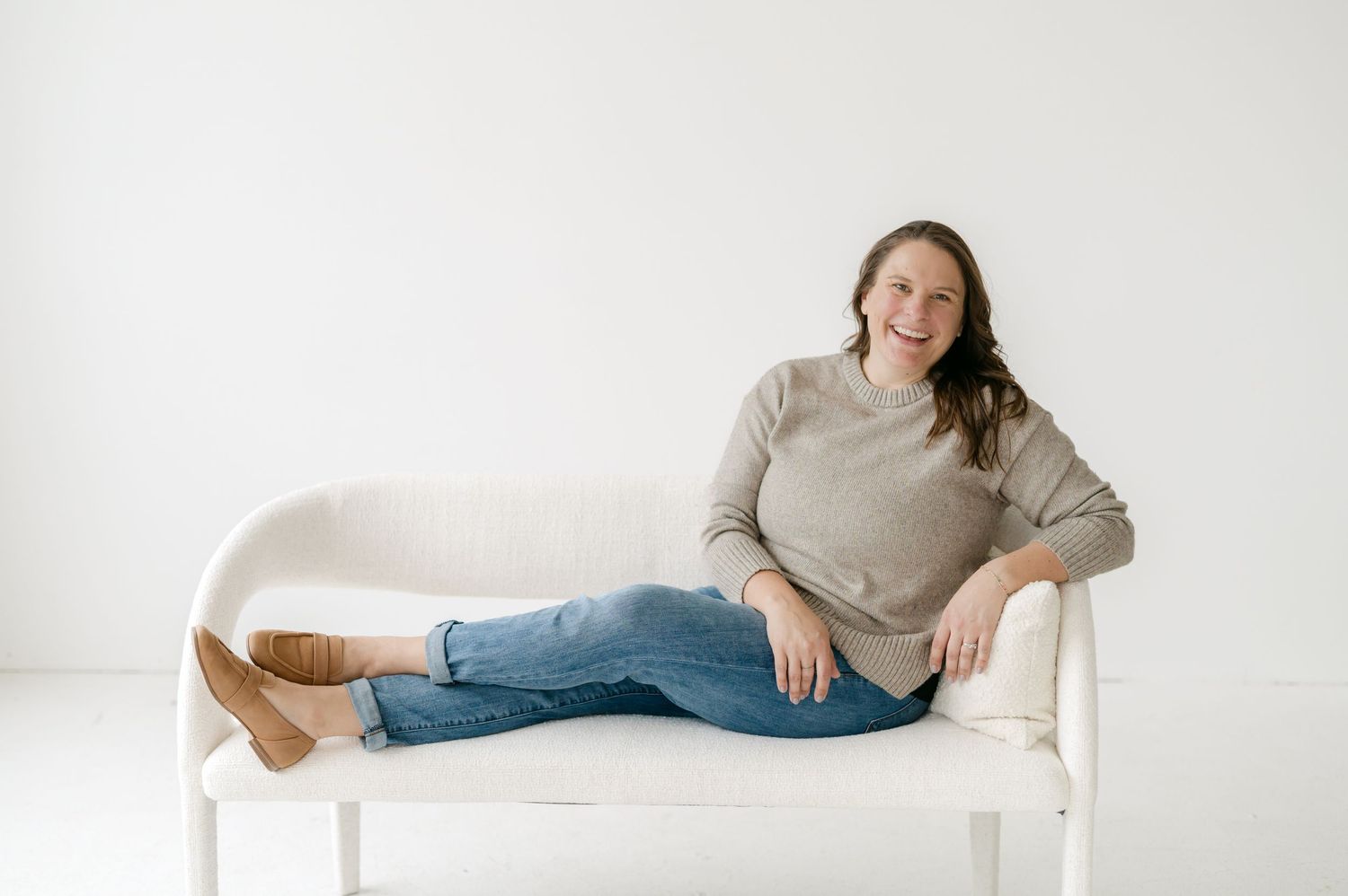 A person wearing a beige sweater and jeans relaxes on a white couch while wearing brown boots.