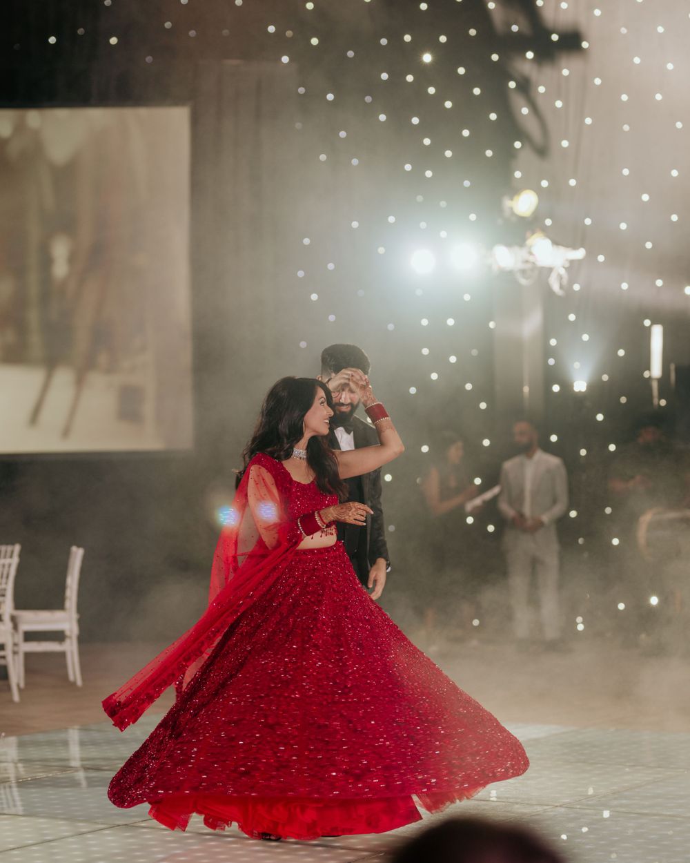 A couple dances elegantly on a misty dance floor, with the woman wearing a stunning red lehenga that twirls with their movement.