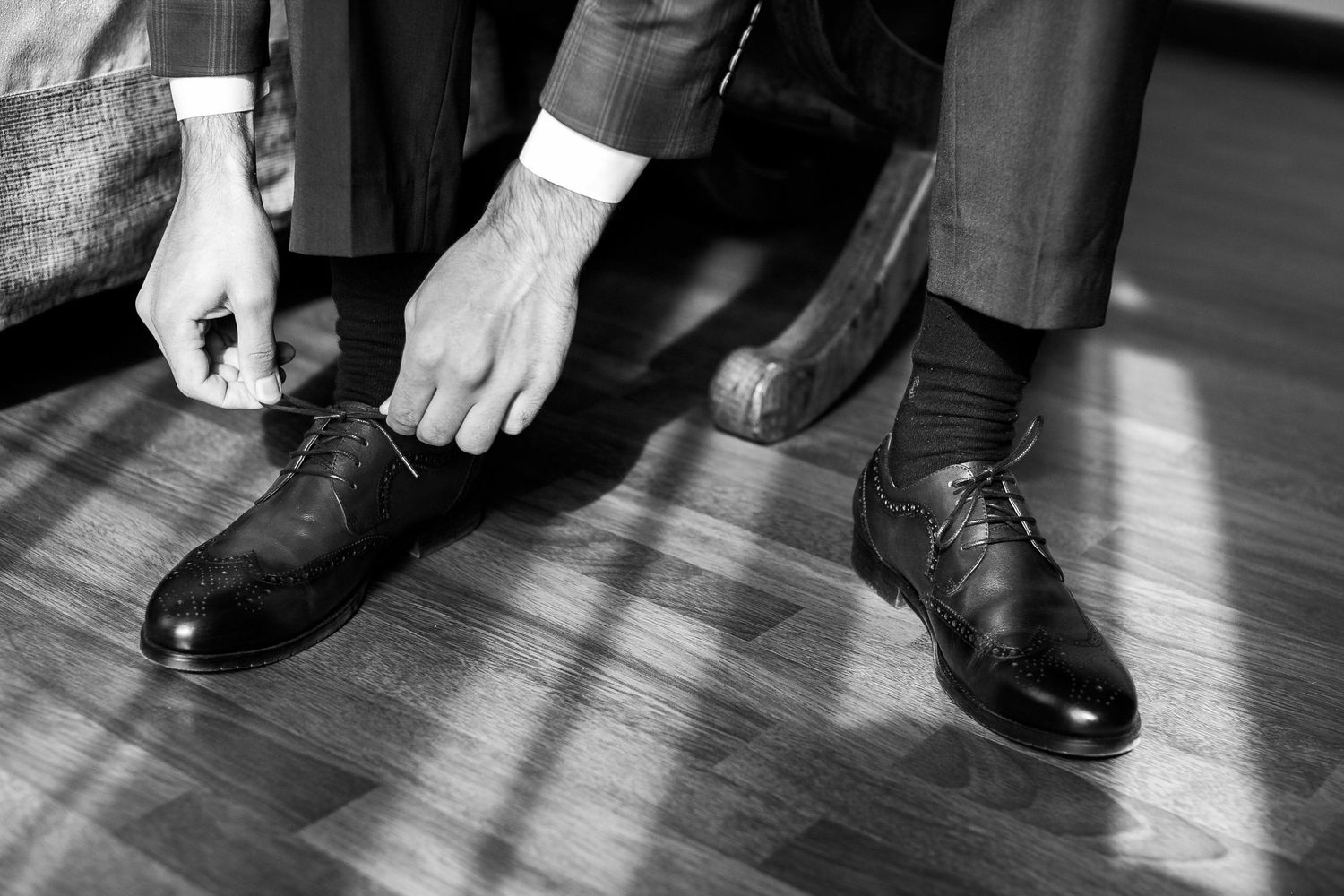 Black and white close-up of hands tying formal dress shoes on hardwood floor.