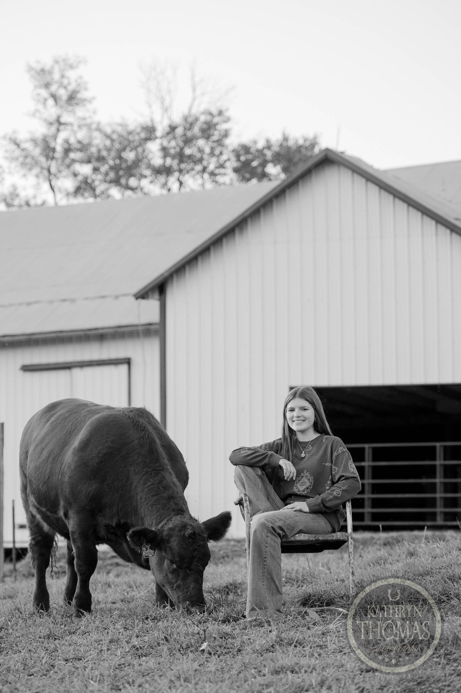 Black and white image of someone seated by a white barn as a cow grazes nearby.
