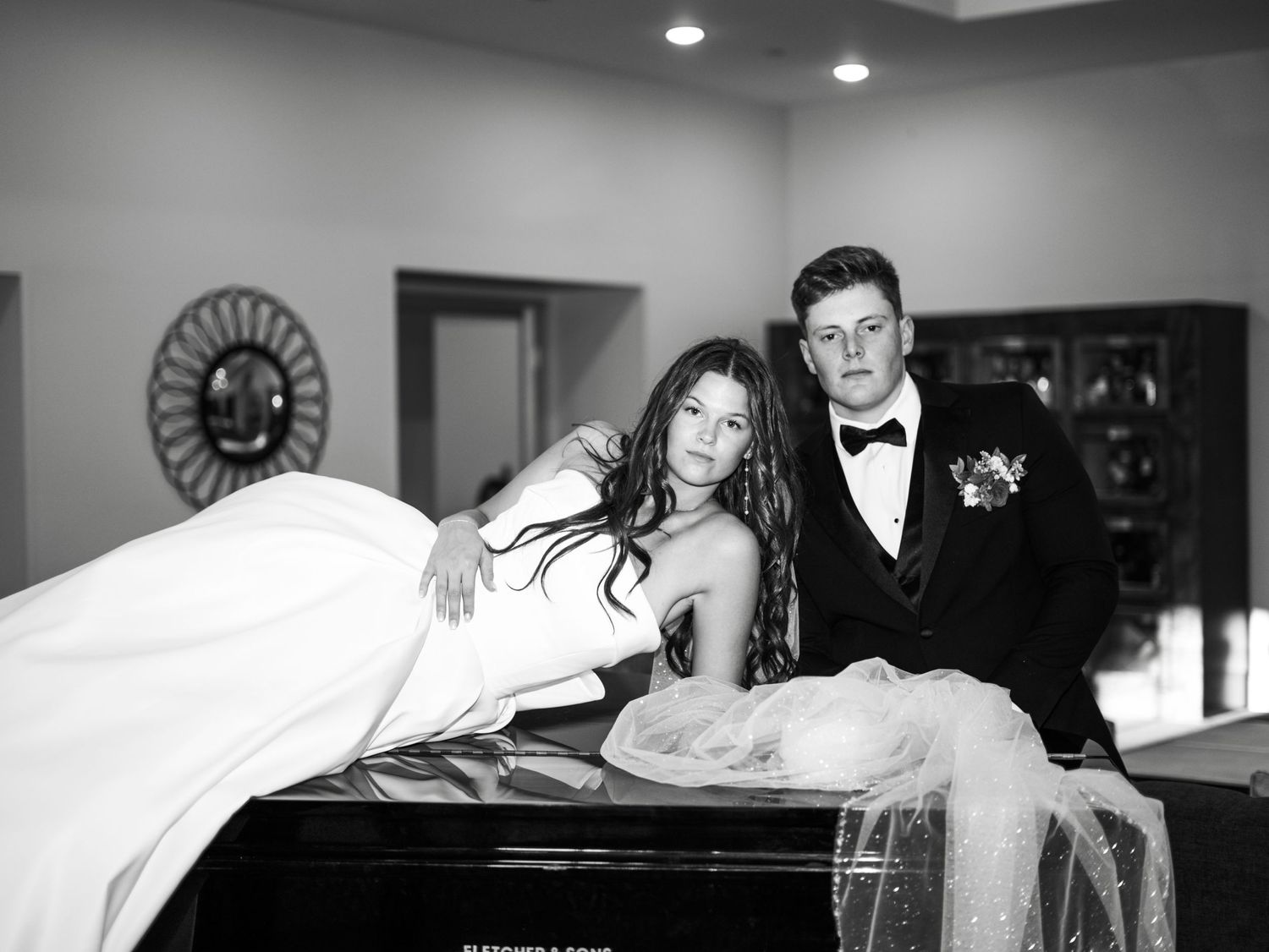 Elegant couple in formal wedding attire pose together in black and white on a piano at their reception celebration.