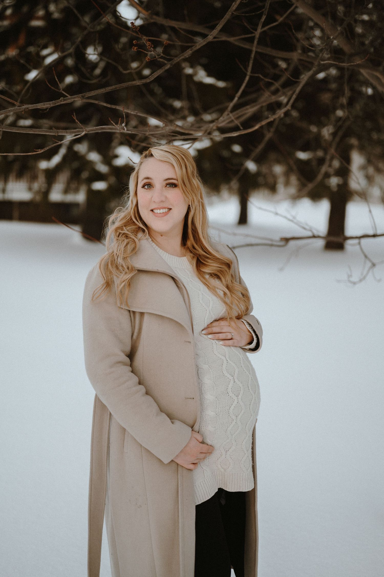 Expectant mother in beige winter coat poses in snowy outdoor setting while cradling her baby bump.