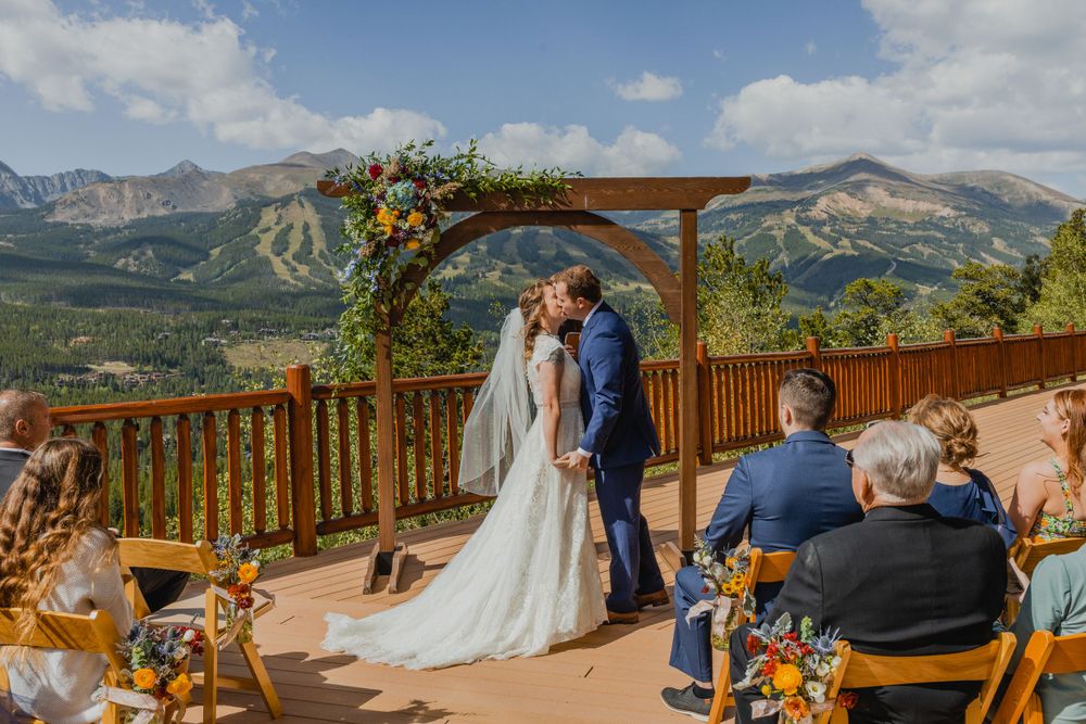 A mountain wedding ceremony takes place on a wooden deck with a floral arch and stunning Colorado peaks in the background.