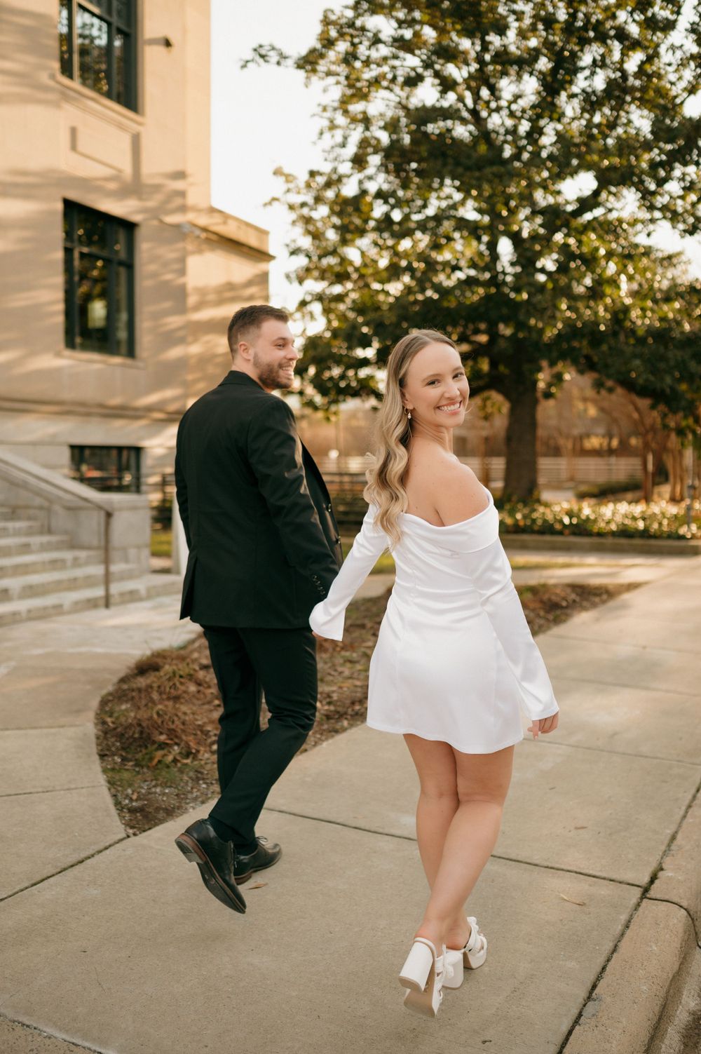 Couple in formal attire walking playfully along sidewalk near modern building.