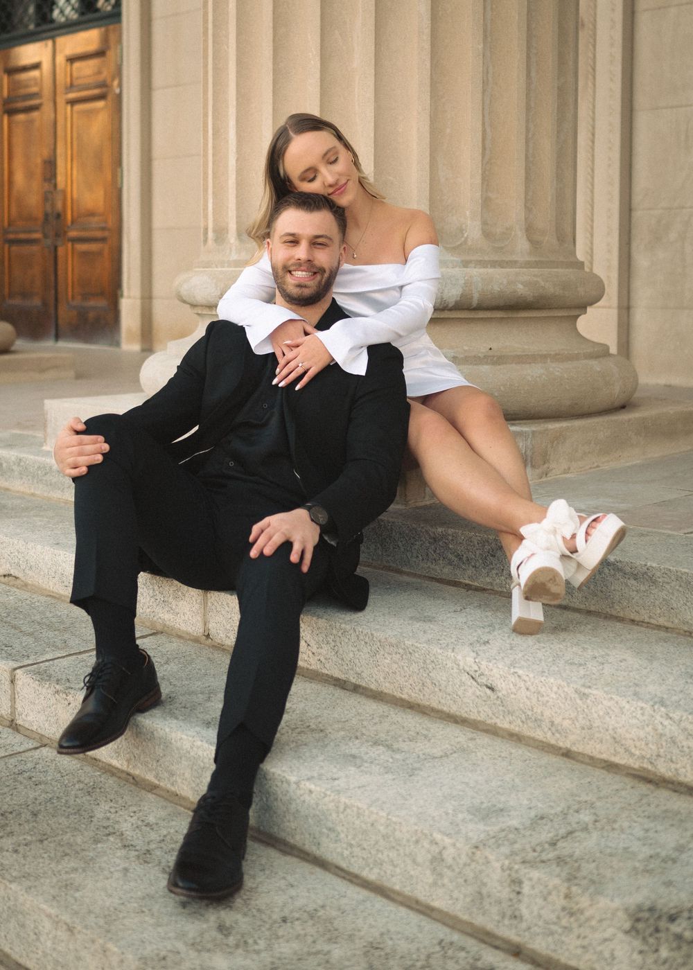 Stylish couple poses casually on stone steps of historic building.