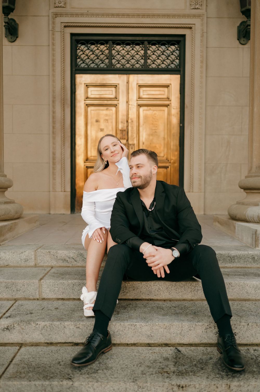 Couple relaxing on stone steps in front of ornate wooden doors.