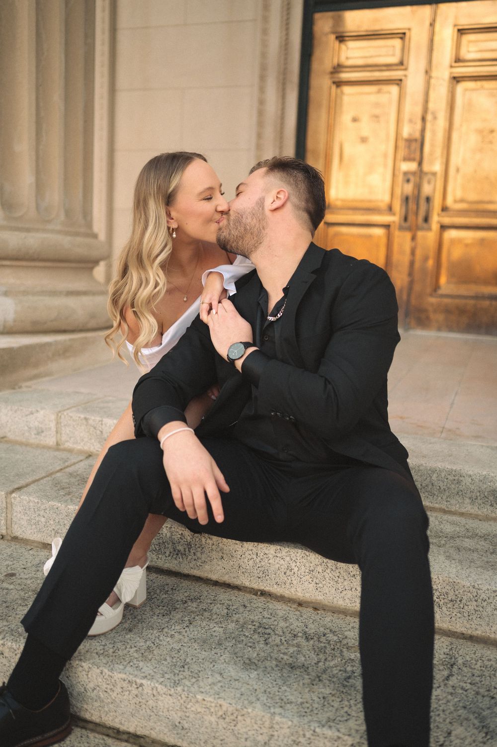 Romantic couple in black formal wear share an intimate moment on stone steps in front of ornate wooden doors.
