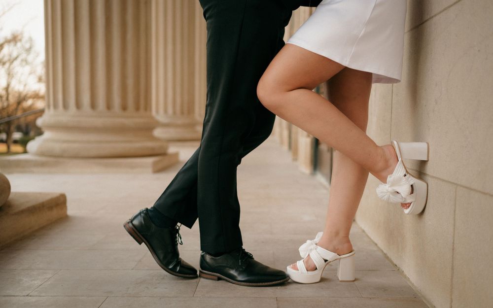 Playful couple's feet detail showing formal black shoes and white heels against classical architecture.