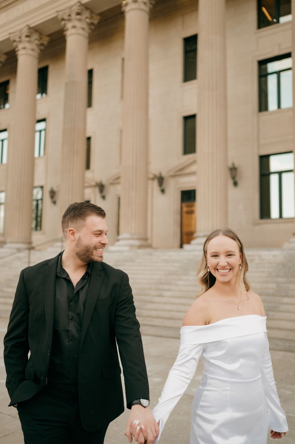 A happy couple in formal wedding attire holding hands while standing on steps of District Attorney office in Charlotte