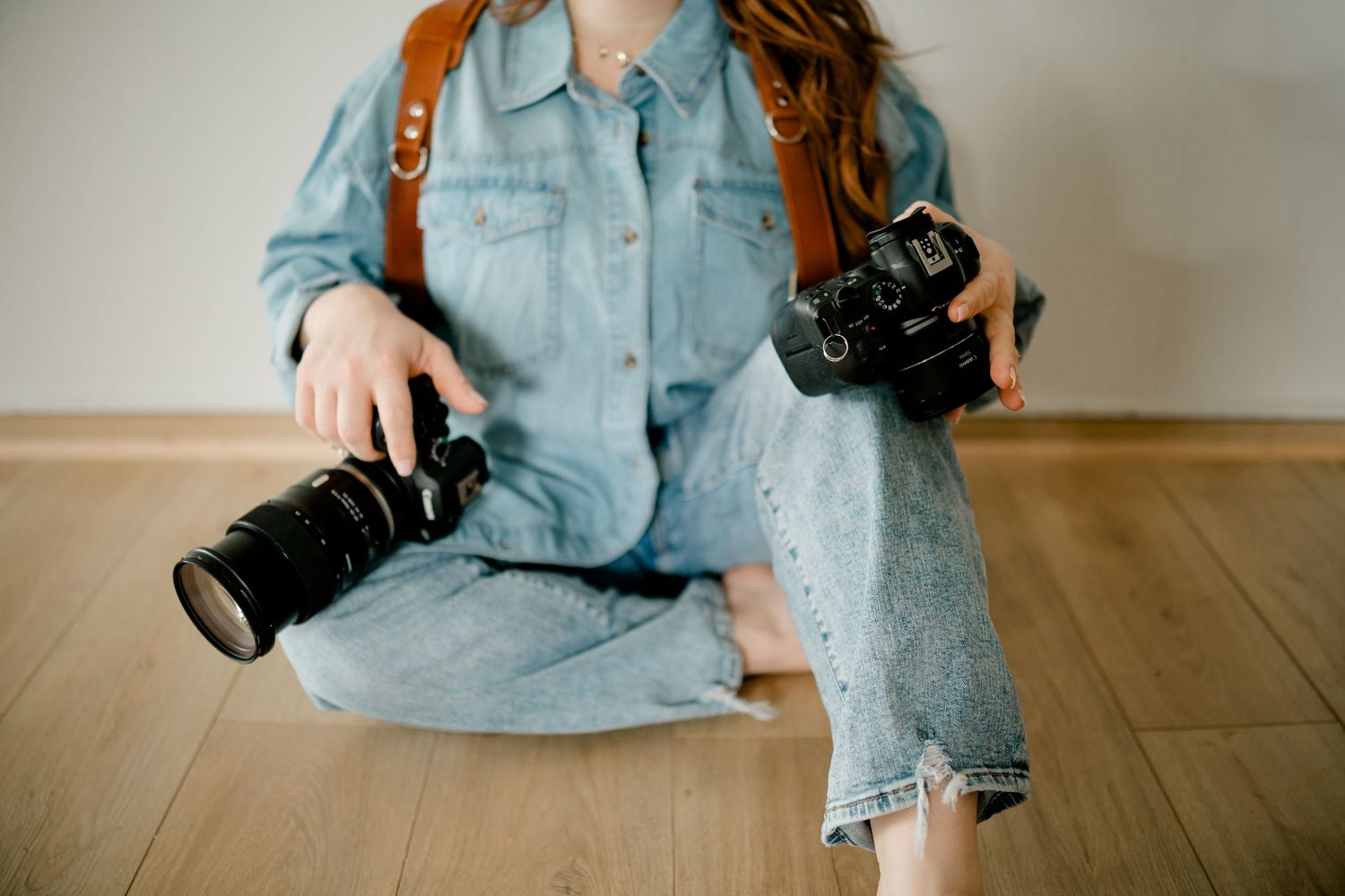 Photographer sitting cross-legged on floor holding two cameras in denim outfit with backpack.