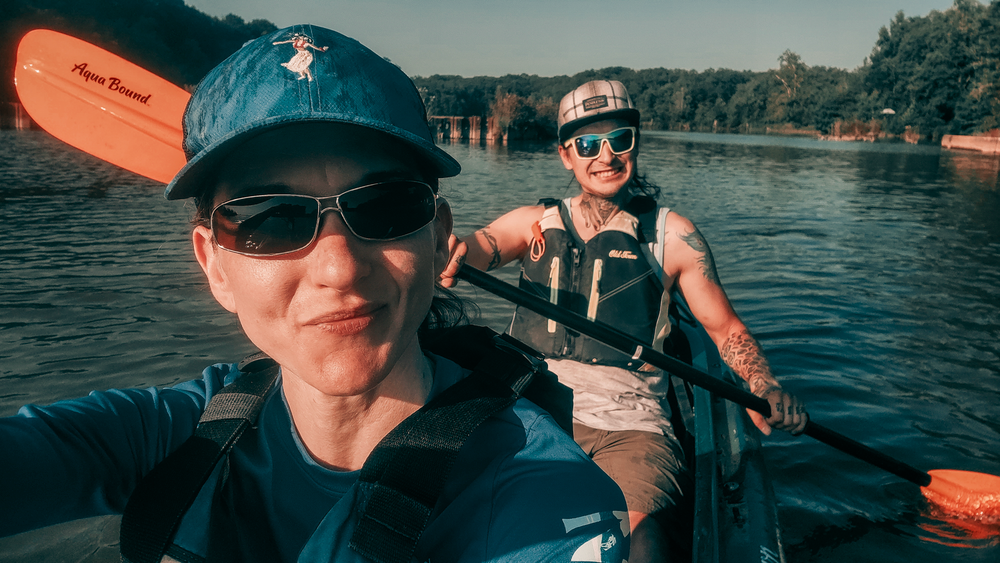 Two kayakers on a lake holding orange paddles while taking a selfie on a sunny summer day.