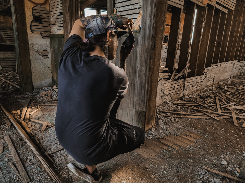 Photographer crouched in dark clothing takes photos inside an abandoned building with exposed wooden beams and debris.