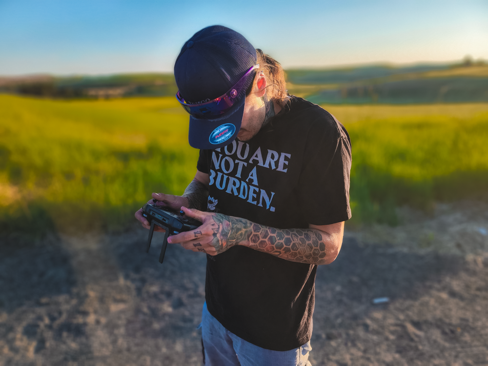 Person in a baseball cap and t-shirt checking phone at sunset in front of a green pastoral field.