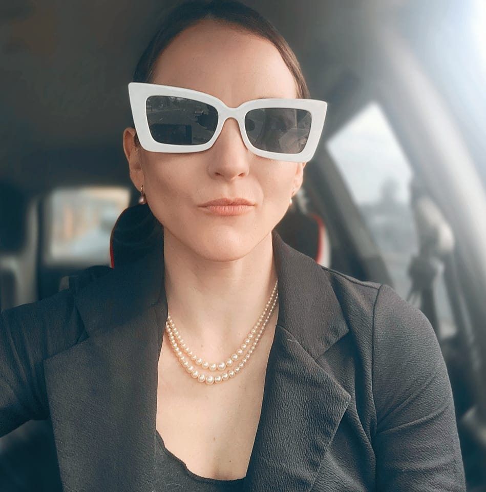 Professional wearing white cat-eye sunglasses, black blazer and pearl necklace sits in car interior.
