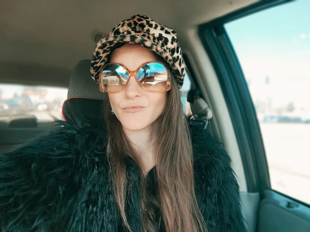 A stylish selfie in a car wearing blue reflective sunglasses, leopard print headwear and a black fuzzy coat.