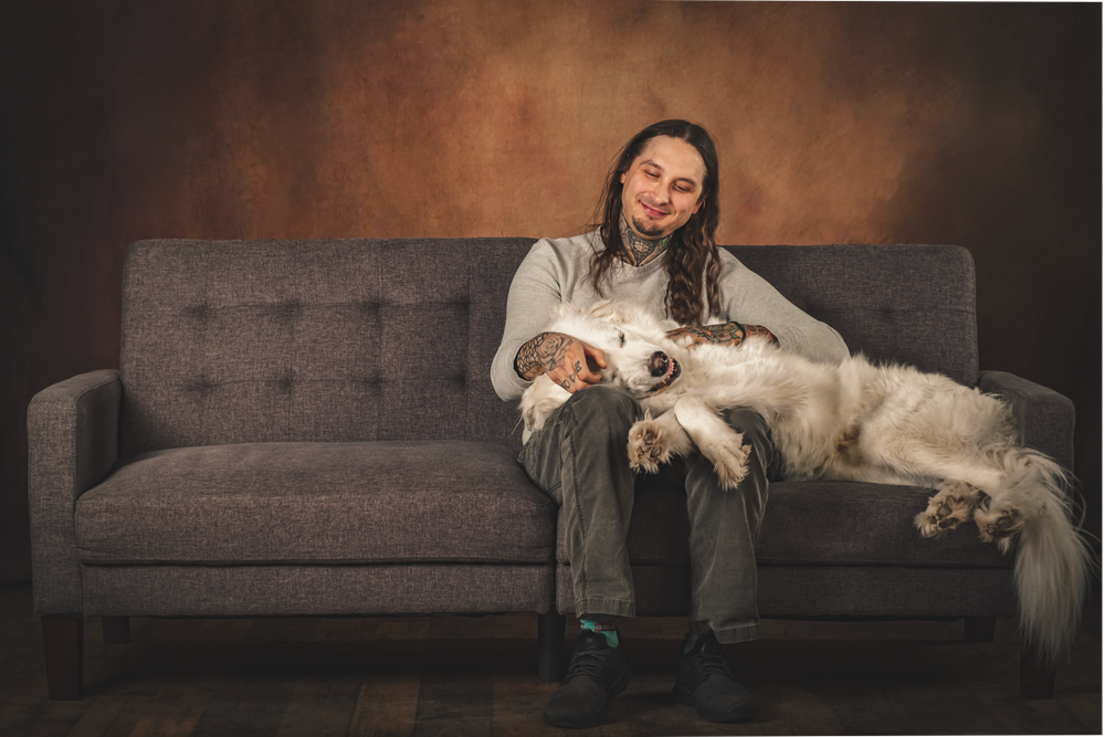 A person sits on a gray couch with a large white fluffy dog lounging next to them in a studio setting.
