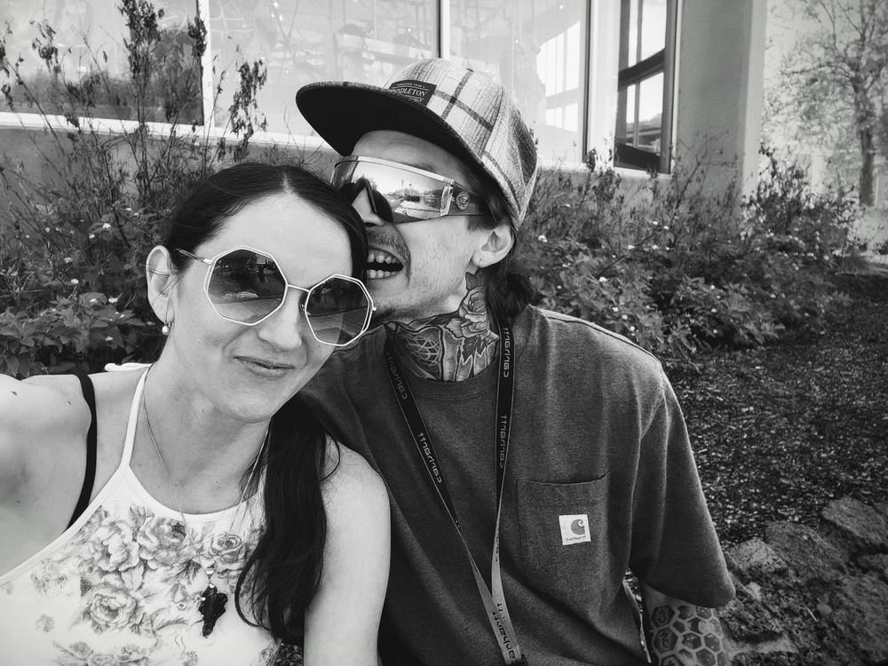 A black and white selfie of a couple outdoors wearing sunglasses and casual attire on a sunny day.