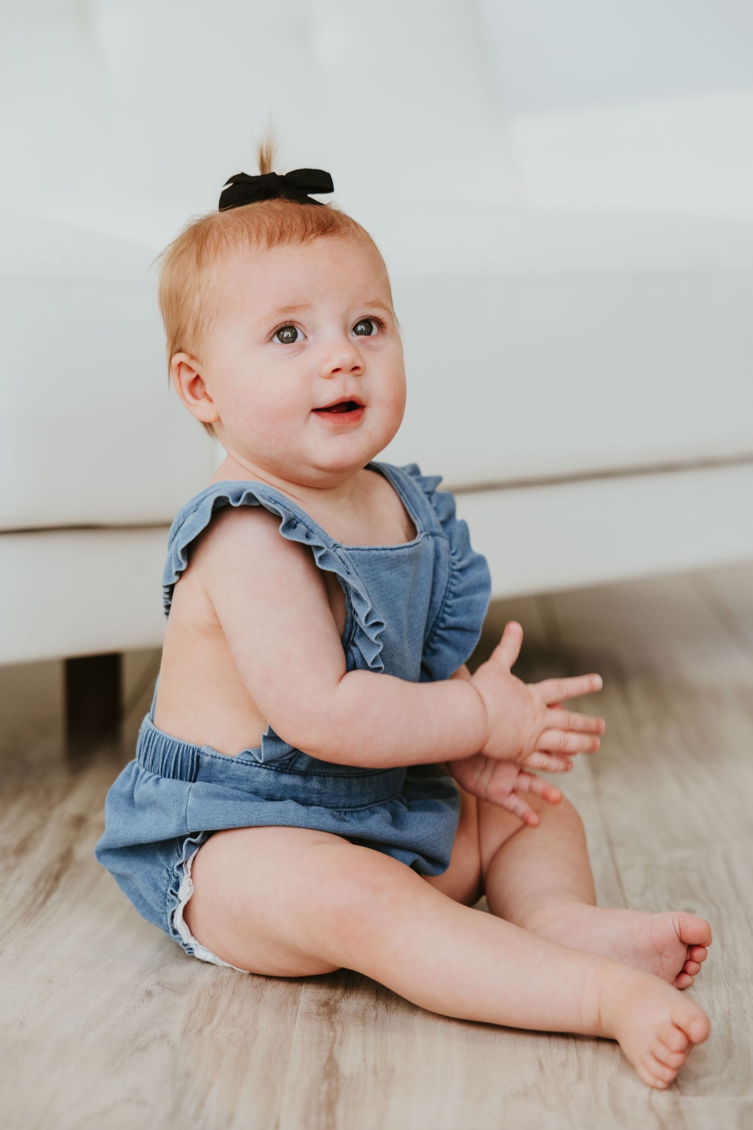 Baby in blue denim romper with black bow sits on wooden floor.