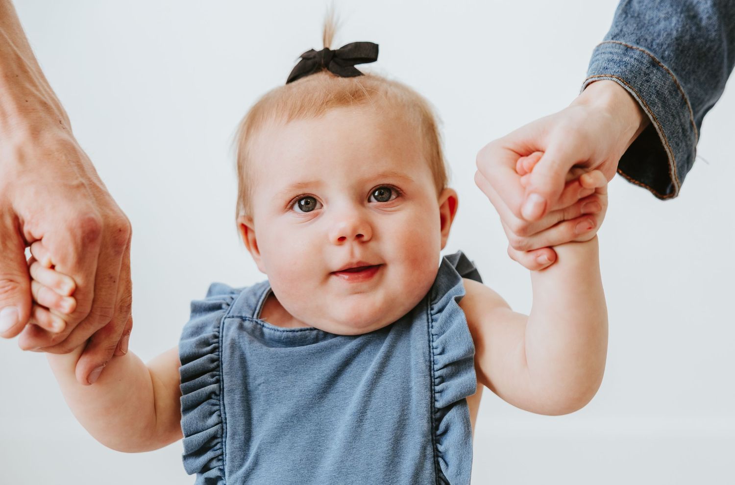 Baby in blue outfit holding hands with adults while standing.