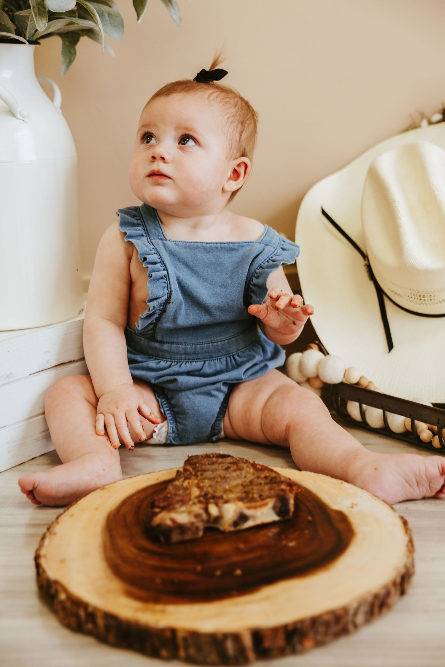 Baby in blue overalls sitting near grilled sandwich on wood slice.