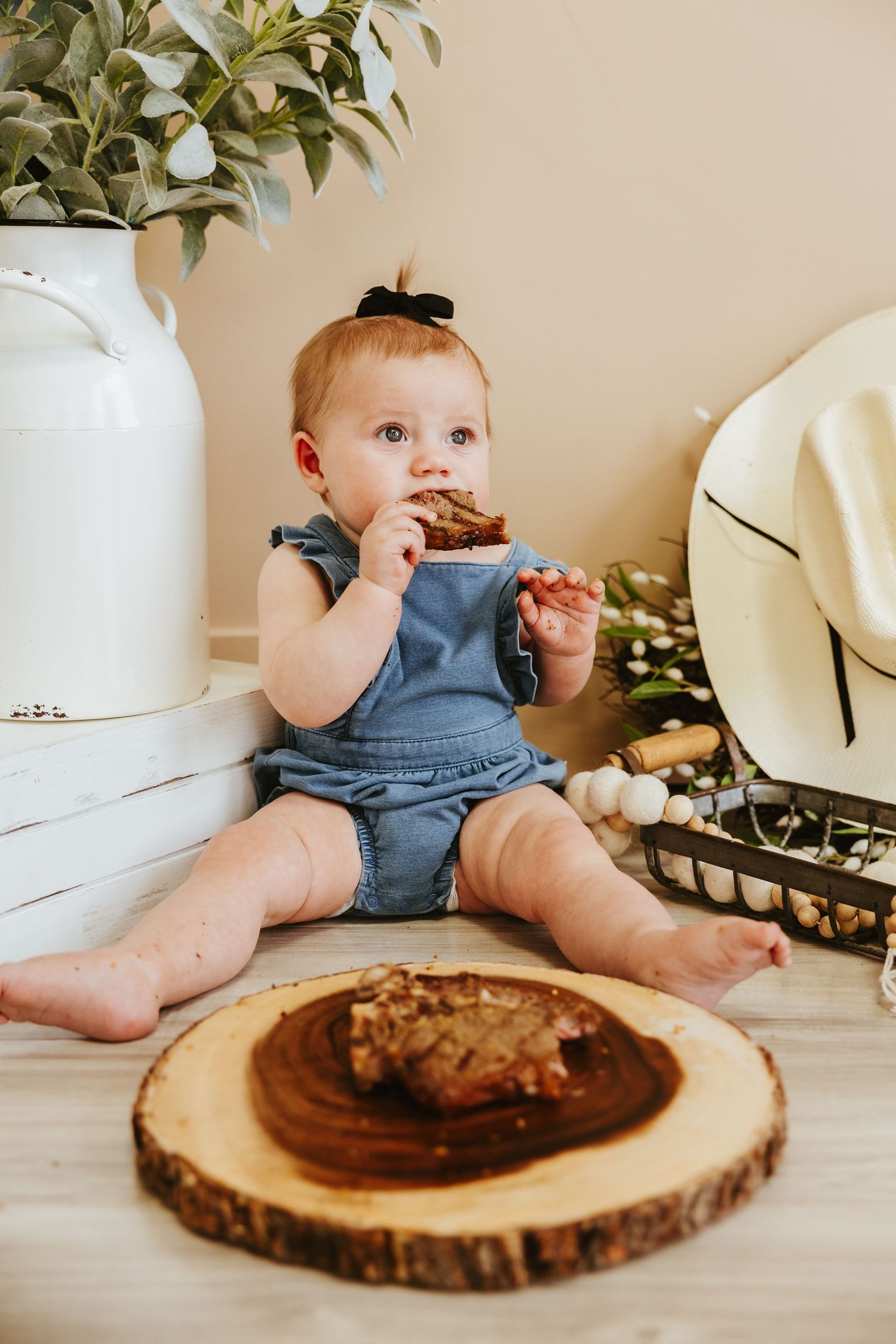 Baby in denim romper enjoying snack while sitting next to white vase.