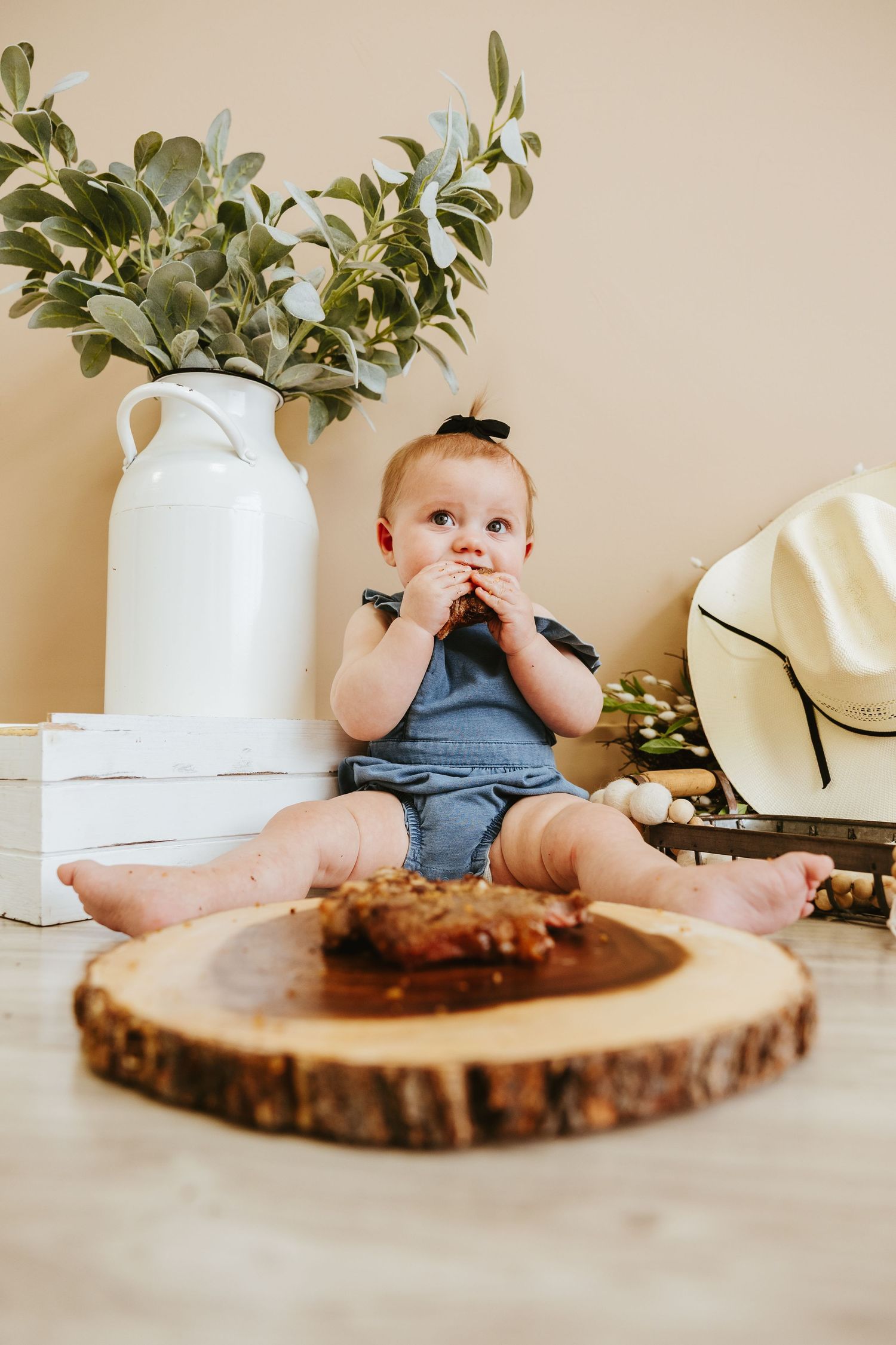 Baby in blue overalls sits near white vase and wooden slice while holding something to their mouth.