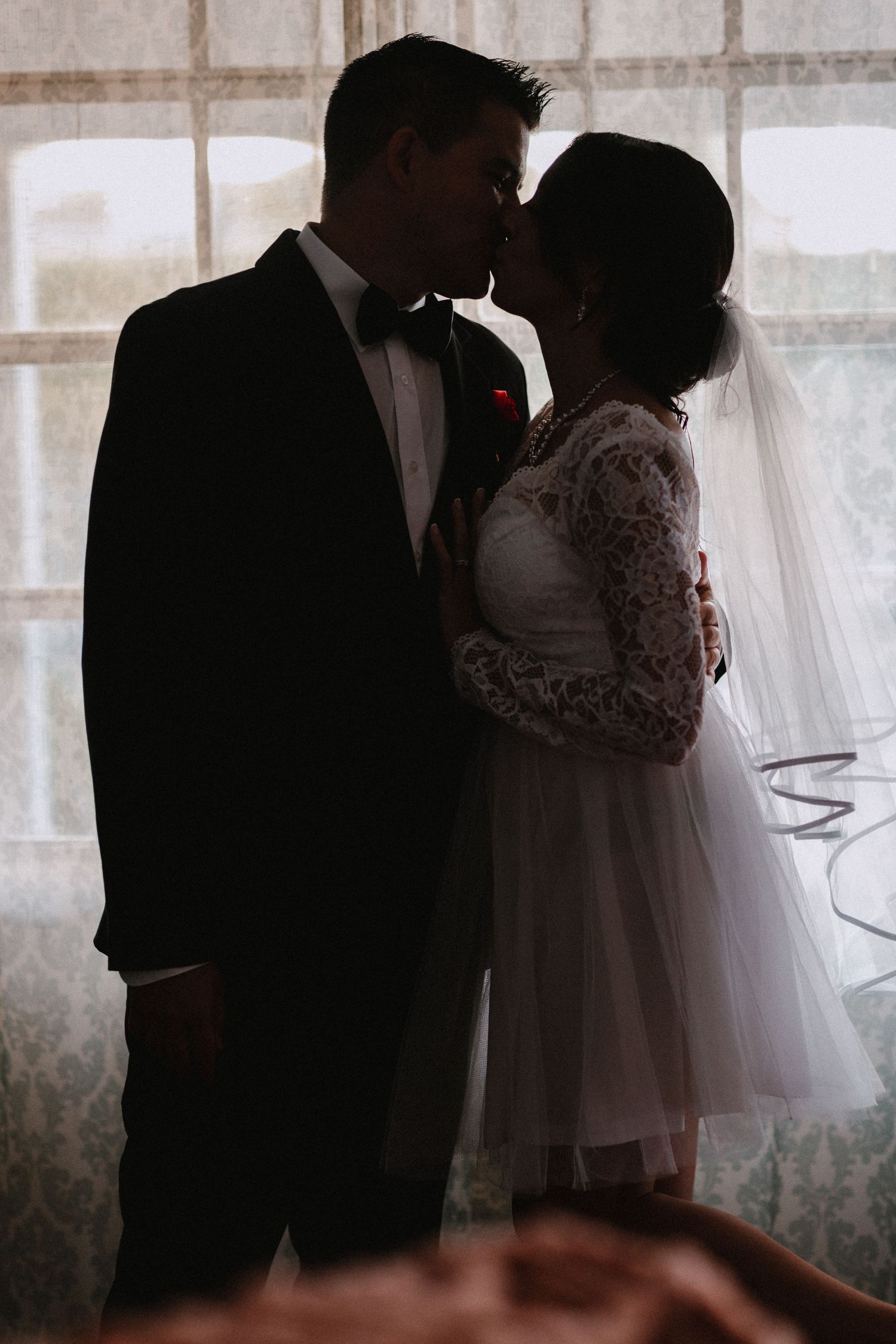 A silhouetted wedding couple share an intimate moment near a window in formal attire.