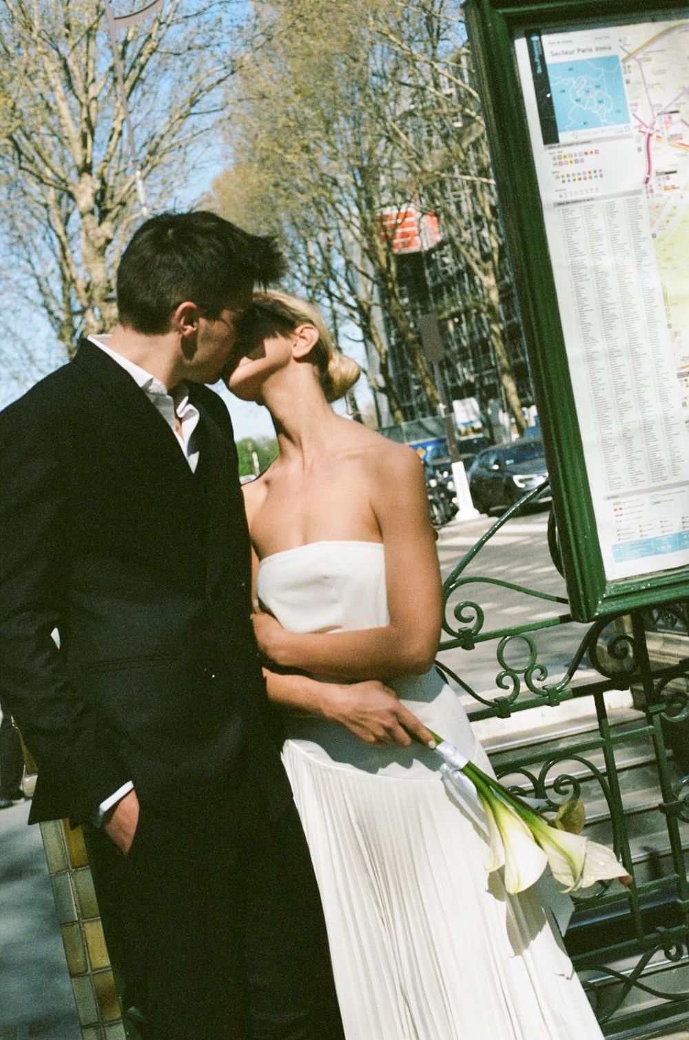 A romantic couple in wedding attire share a kiss by a city map display on a sunny day.