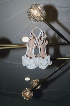 Delicate white bridal shoes with lace details displayed against a dark ceiling with lighting fixtures.
