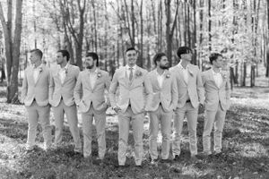 Groomsmen in light colored suits lined up for a formal wedding photo in a wooded setting.