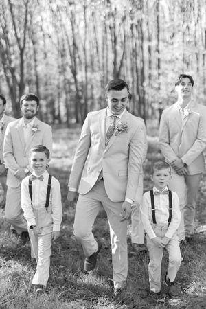 Wedding party with young ring bearers in suspenders and light colored suits posing in a forest setting.