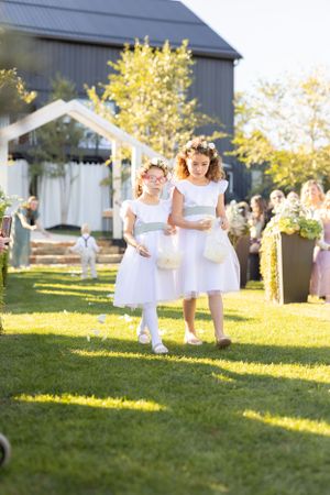 Two flower girls in white dresses running on a sunny lawn during an outdoor wedding ceremony at a rustic barn venue.